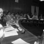 A black-and-white image of Dr. Manmohan Singh seated at a conference table with government officials. Holding documents, he appears focused during a high-level economic discussion. Other officials, including economists and policymakers, are also present in the room.