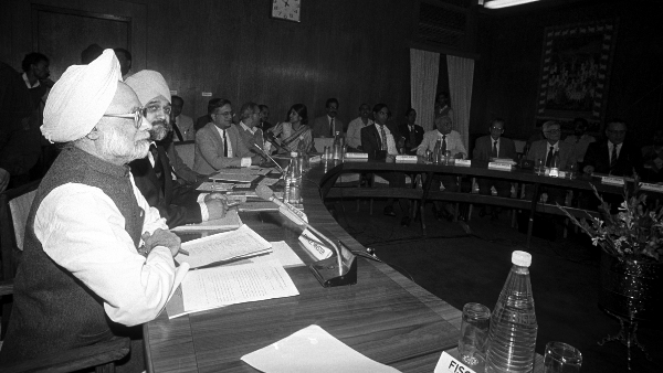 A black-and-white image of Dr. Manmohan Singh seated at a conference table with government officials. Holding documents, he appears focused during a high-level economic discussion. Other officials, including economists and policymakers, are also present in the room.